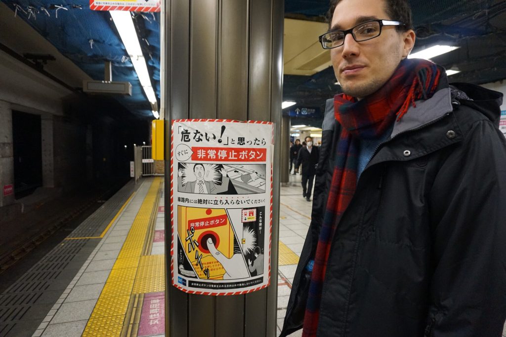 Greg from You Could Travel in front of a sign in Japan telling people to take action if someone falls on the subway tracks