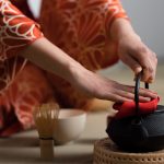 a Japanese woman dressed in a silk kimono preparing to pour hot water from a kettle for the tea ceremony